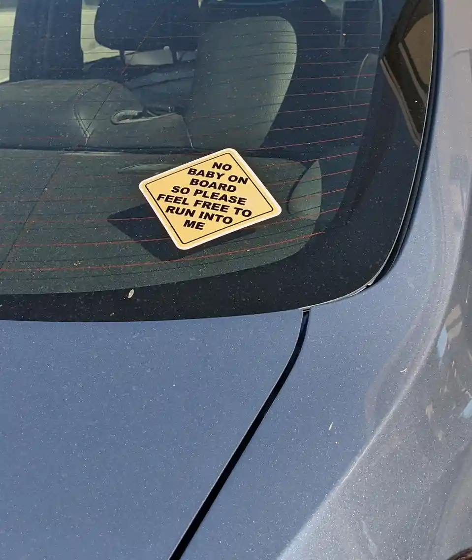 A yellow diamond-shaped sticker on a car's rear window reads 'NO BABY ON BOARD SO PLEASE FEEL FREE TO RUN INTO ME'…