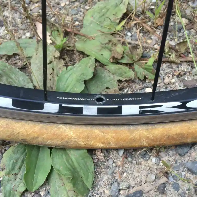 A close-up photo shows a black bicycle wheel rim with the text 'ALUMINIUM ALL ETRTO 622X15C' printed on it, lying on gravel…