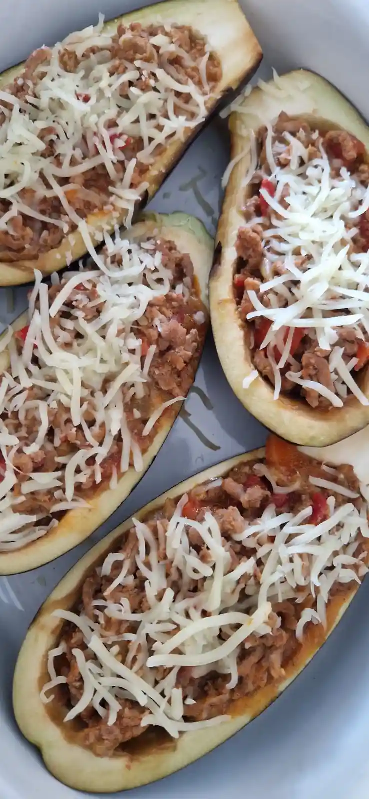 Four halved eggplants are arranged in a baking dish, each filled with a meaty sauce and topped with shredded white cheese…