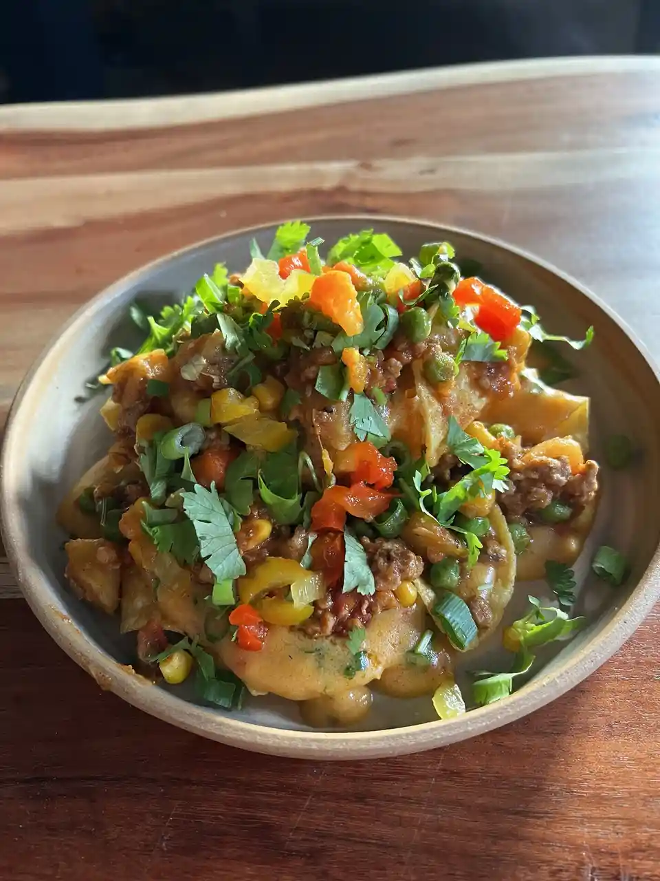 A close-up photo shows a bowl of loaded nachos on a wooden table, topped with melted cheese, ground meat, chopped green…