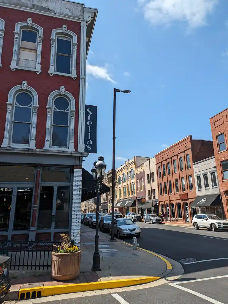 A sunny street view of a historic downtown with red brick buildings, parked cars, and a prominent black vertical sign…