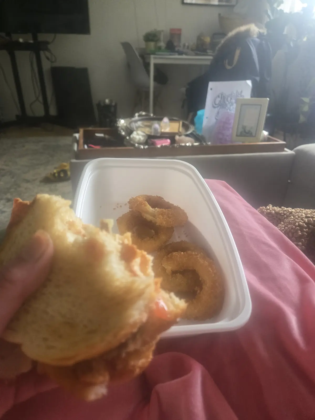 A person holds a partially eaten sandwich in front of a white takeout container filled with fried onion rings, resting on a…
