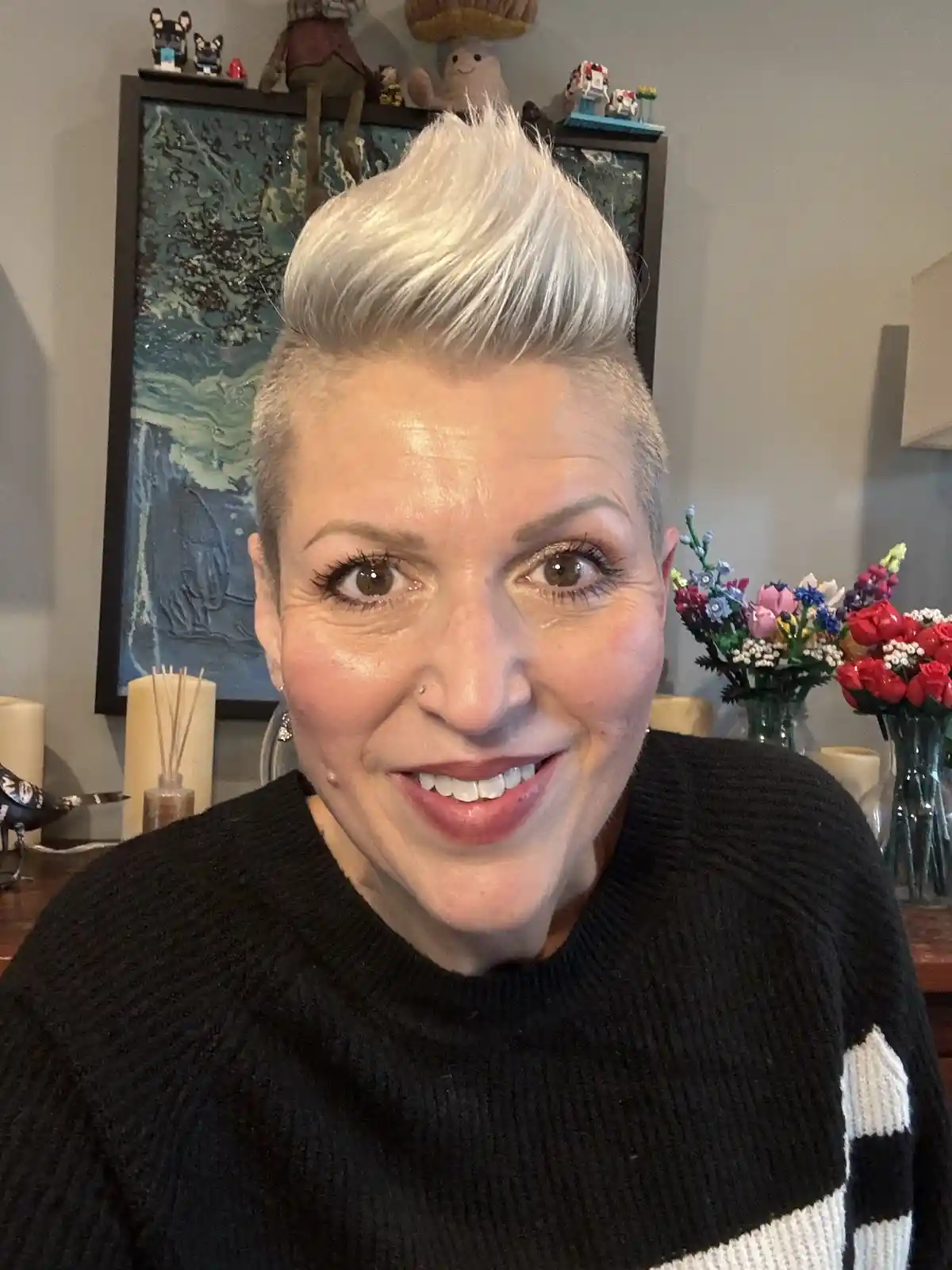 A woman with short, platinum blonde hair styled in a high, spiky mohawk haircut smiles at the camera while wearing a black…
