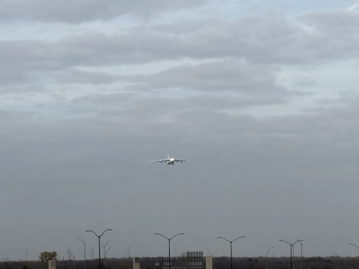 A white passenger airplane is captured mid-flight against a cloudy, overcast sky, flying low over a distant horizon with…