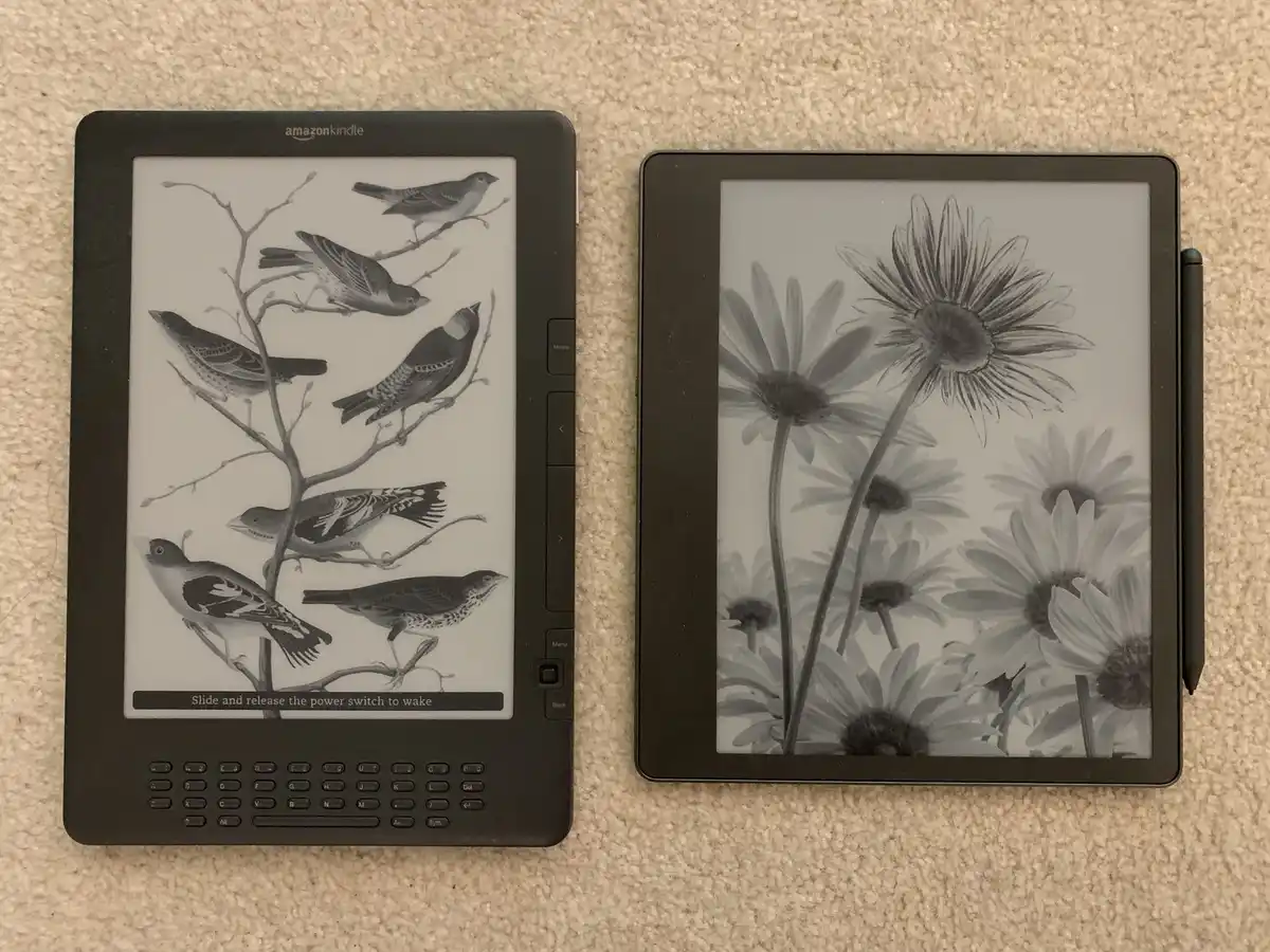 Two black-and-white e-readers lie side-by-side on a beige carpet. The left device, an older Amazon Kindle with a physical…
