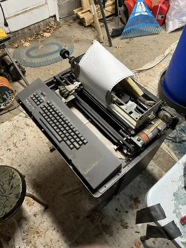 A modified, open-frame typewriter with exposed internal mechanisms and a keyboard, sitting on a concrete floor surrounded by…