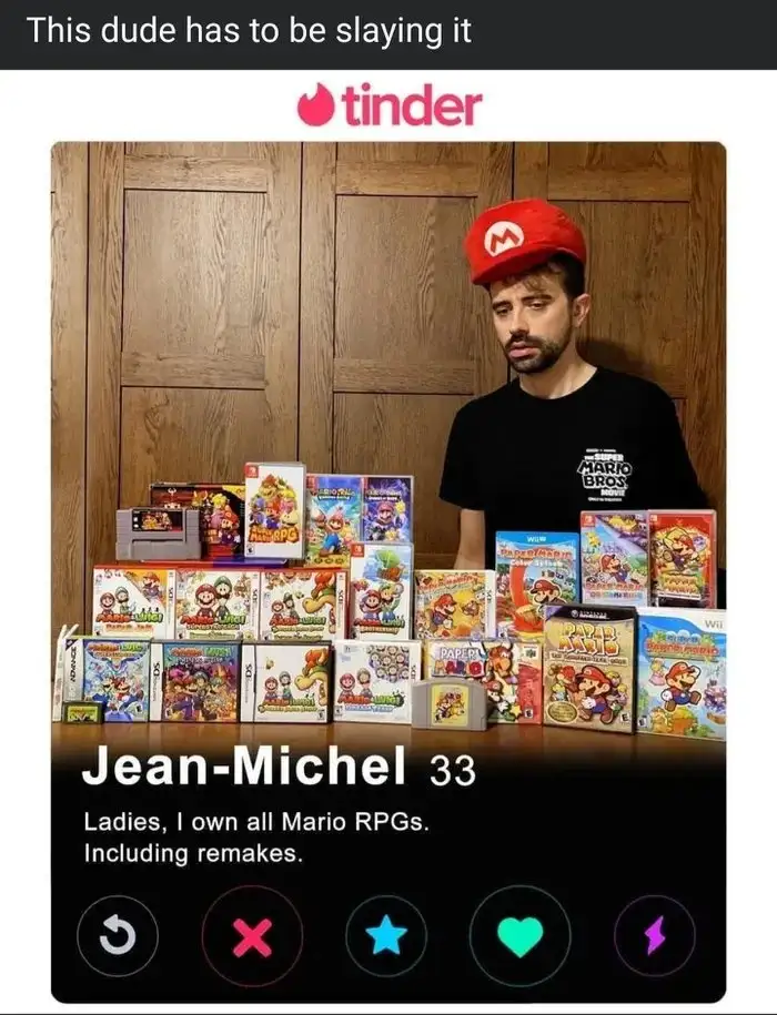A man wearing a red Mario cap and a black t-shirt stands behind a large collection of Mario RPG video game boxes arranged…