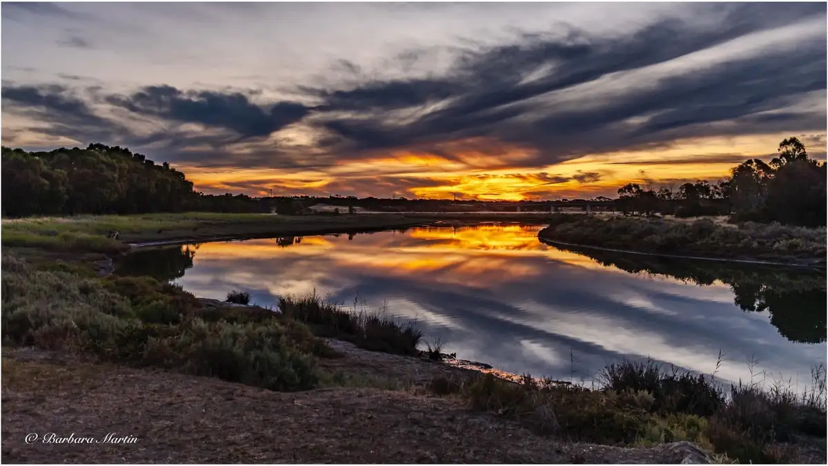 A serene landscape photograph captures a river reflecting a dramatic sunset with orange and dark clouds, flanked by grassy…