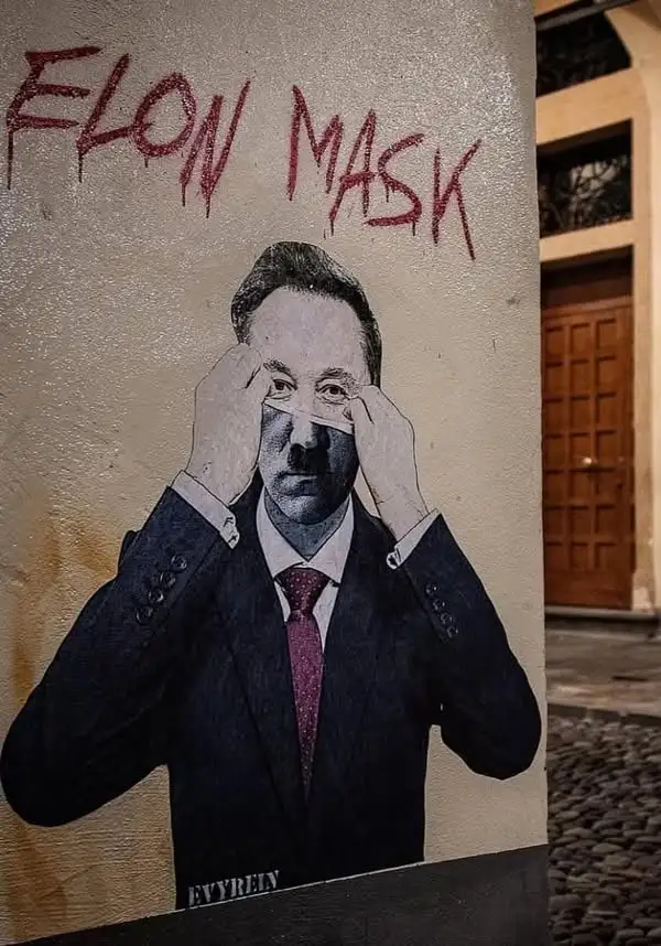 A street mural depicts a man in a suit and tie, pulling a white strip of tape across his eyes, with the words 'ELON MASK'…