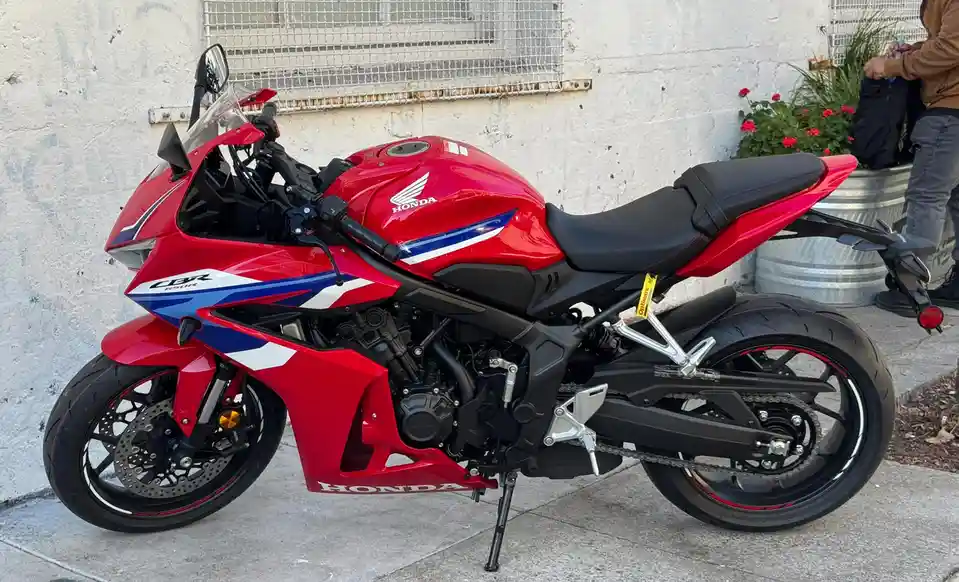 A red Honda CBR650R sport motorcycle with blue and white accents is parked on a sidewalk in front of a white wall with a…