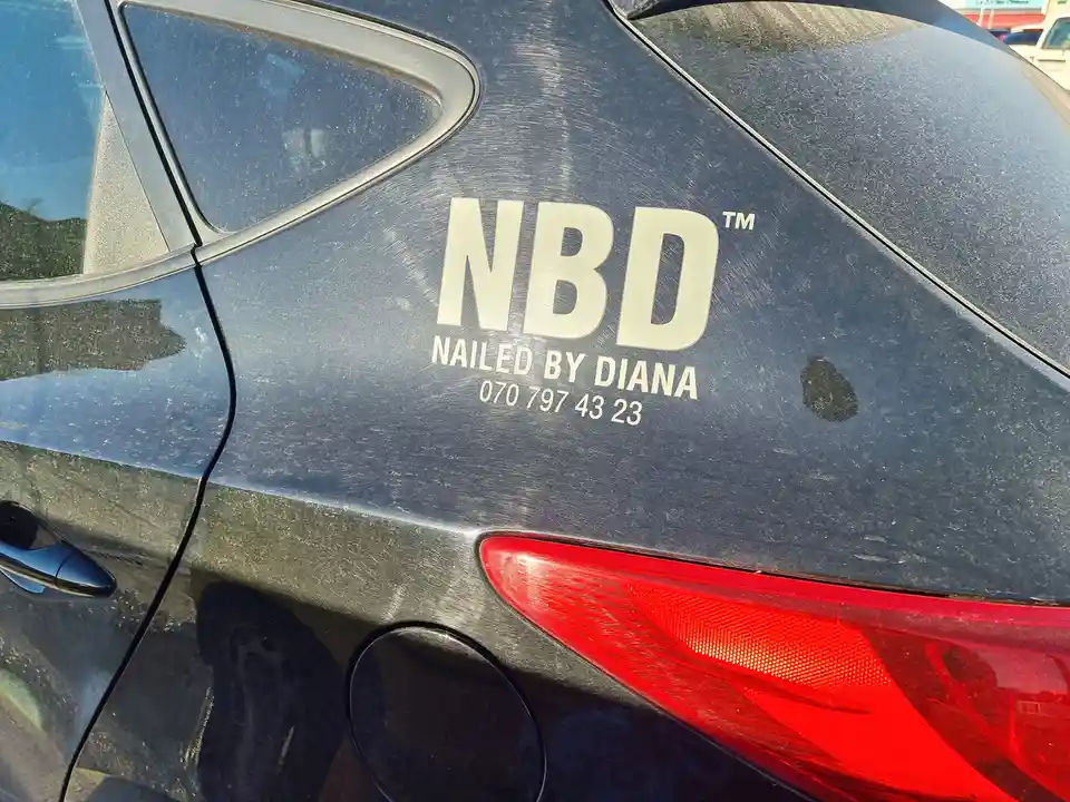 A close-up of a dark-colored car's rear door shows a custom decal with the text 'NBD™ NAILED BY DIANA 070 797 43 23'. The…