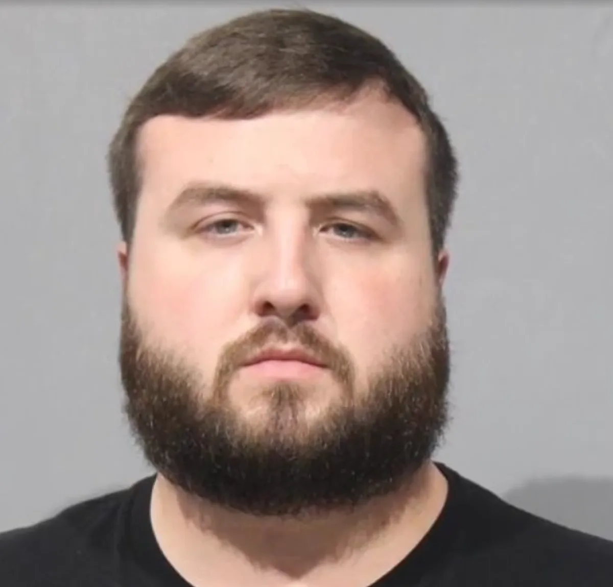 A mugshot-style photo of a man with a full beard and short brown hair, wearing a black t-shirt, looking directly at the…
