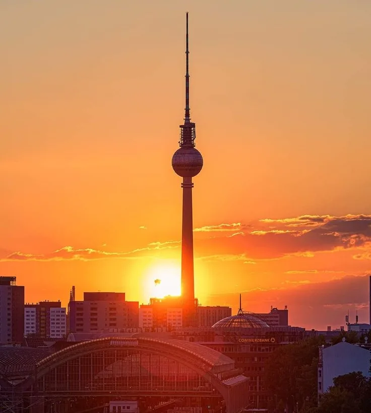 A scenic sunset photo of the Berlin TV Tower (Fernsehturm) silhouetted against an orange sky with the sun low on the…