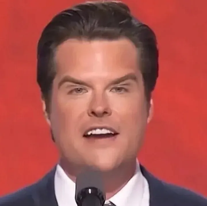 A close-up of a man with dark hair and a suit speaking into a microphone against a solid red background. His expression is…