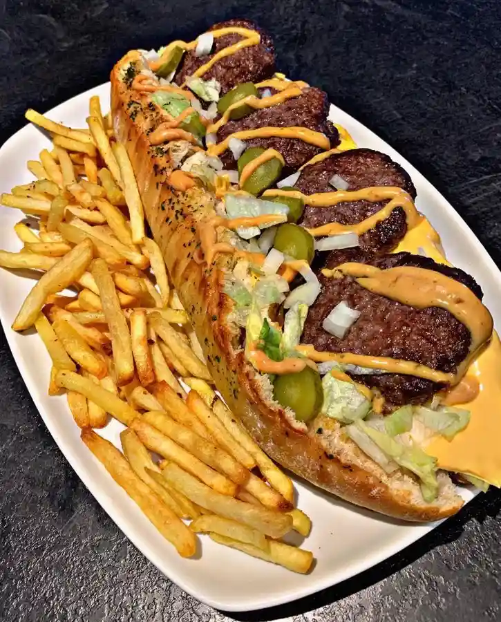 a close-up photo of a long hot dog bun filled with multiple grilled beef patties, melted cheese, pickles, onions, and a…