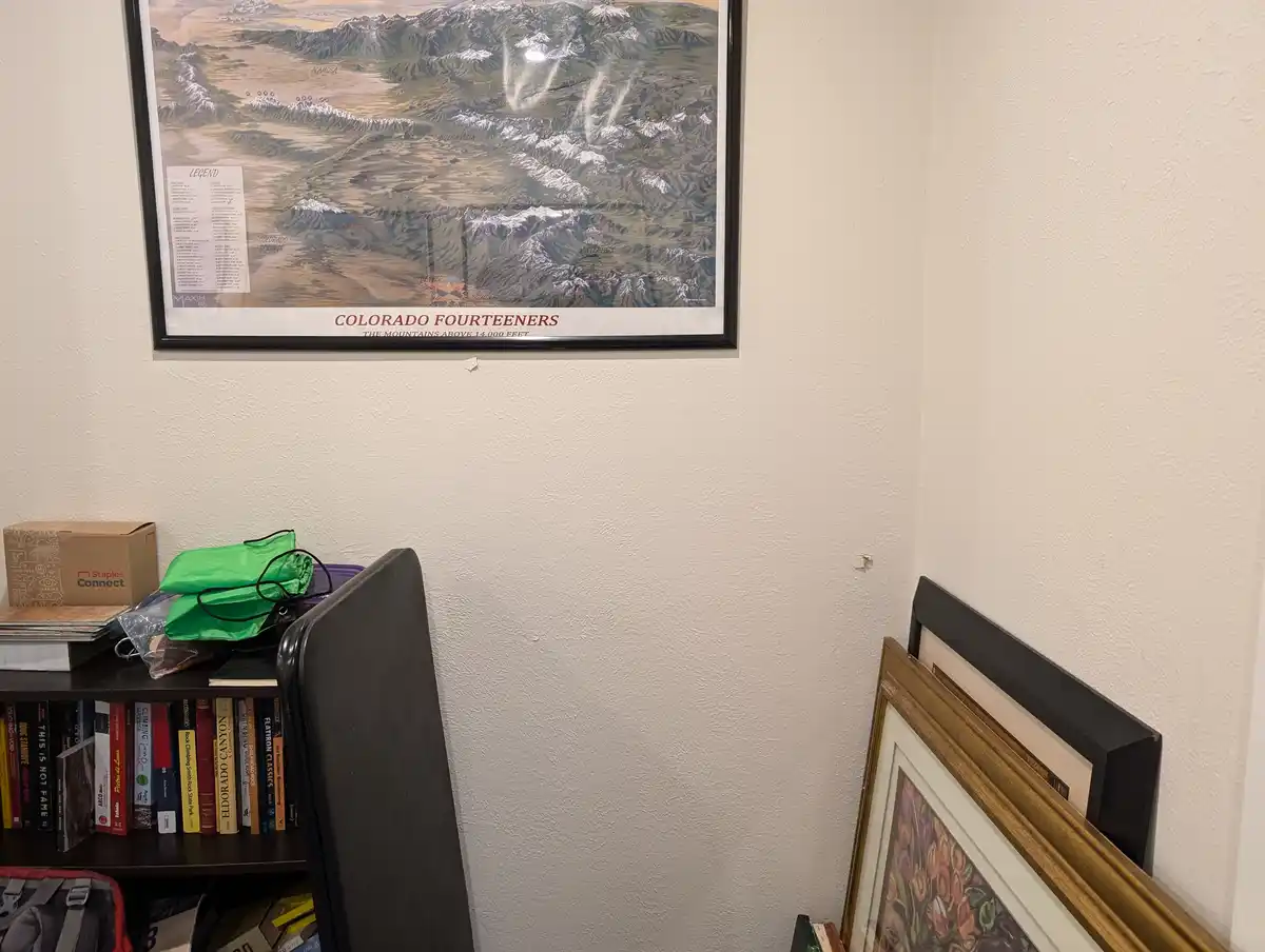 A framed map of Colorado Fourteeners hangs on a wall above a bookshelf; the map shows mountain ranges and peaks with a…
