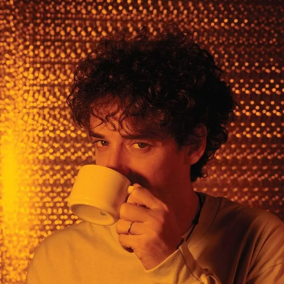 A person with curly dark hair holds a white mug to their mouth, partially obscuring their face, against a warm-toned…