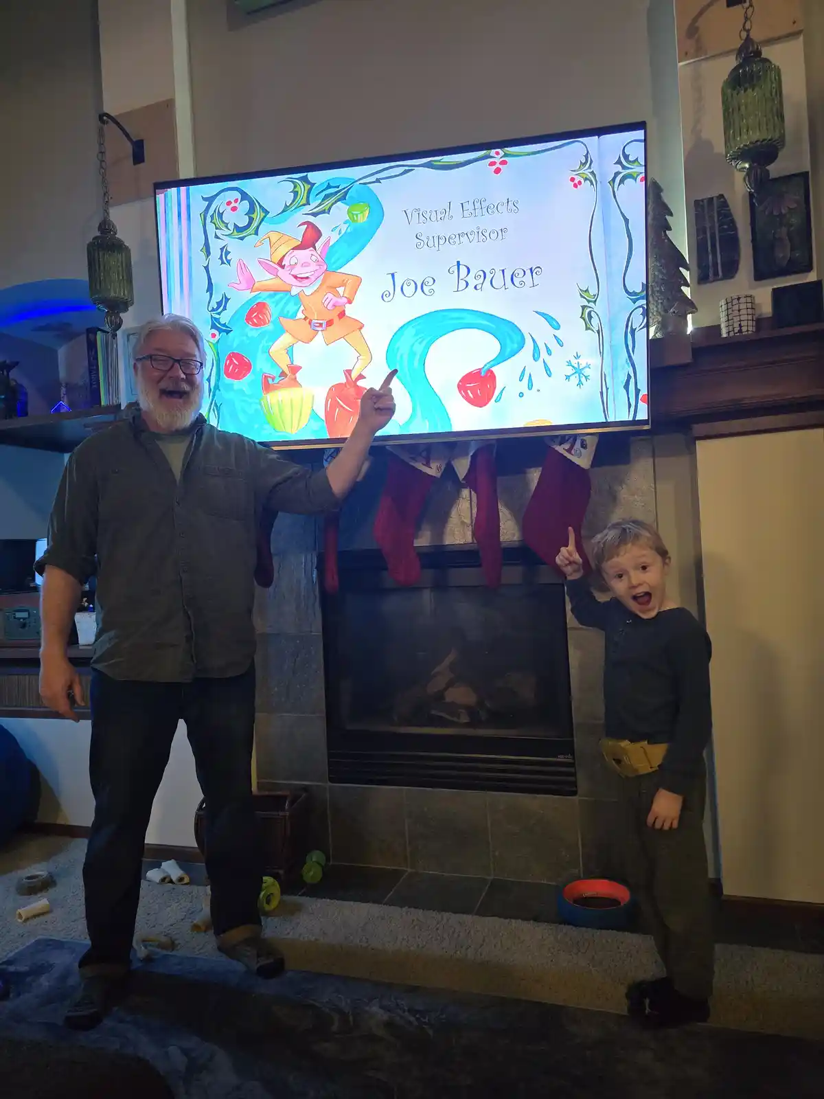 A man and a young boy stand in front of a fireplace, both pointing excitedly at a TV screen displaying a colorful animated…
