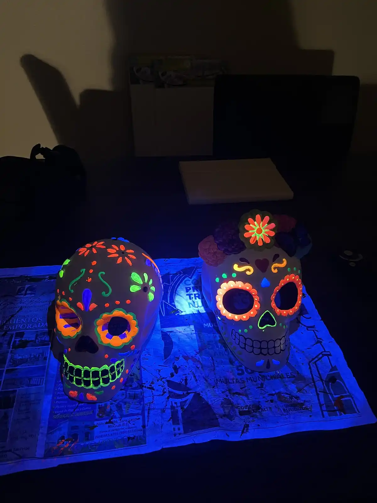Two decorated sugar skulls, glowing brightly under blacklight, rest on a newspaper on a dark desk. The skulls are painted…