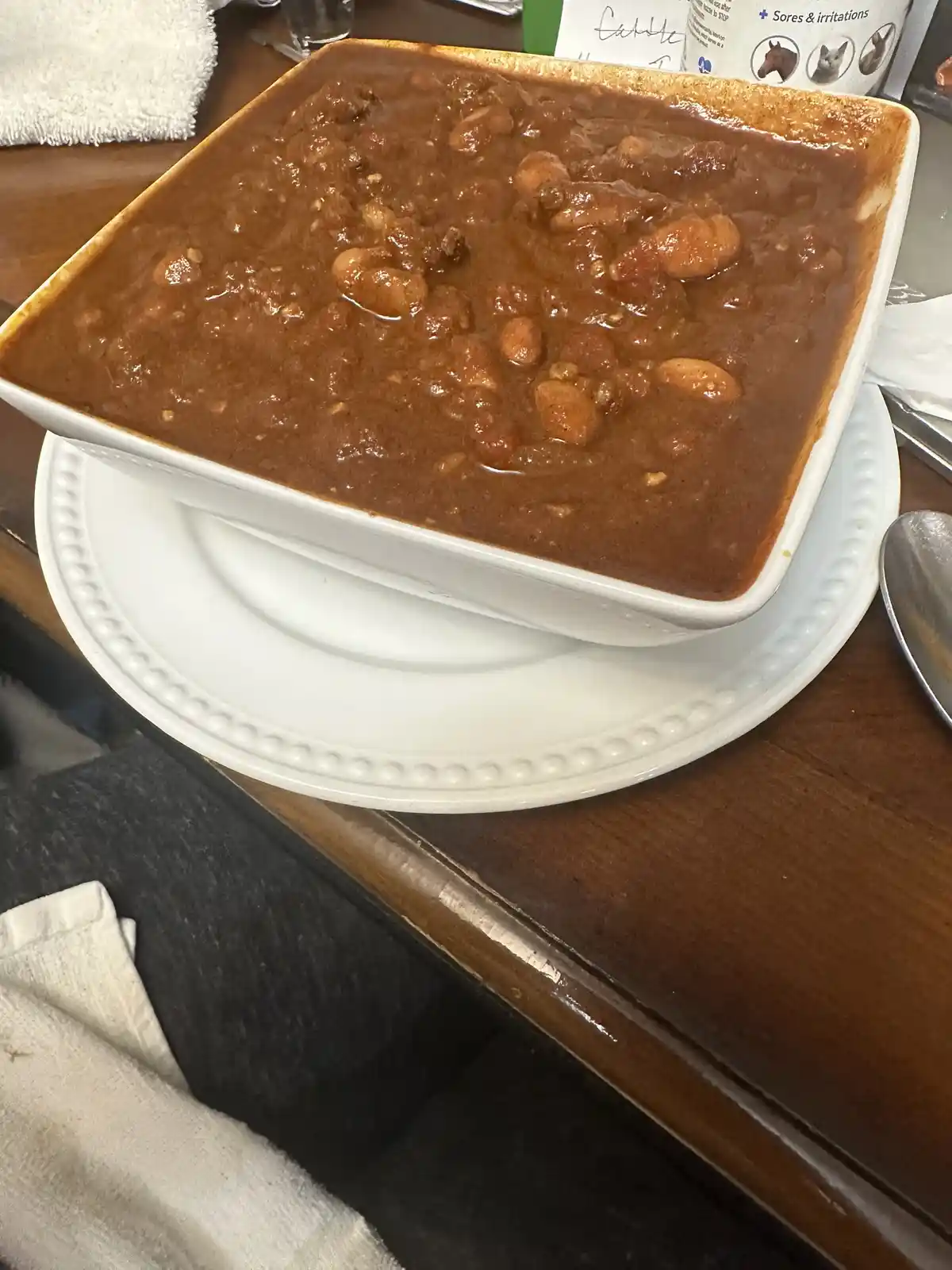 A square white ceramic bowl filled with a thick, brown, bean-heavy stew sits on a white plate atop a wooden table. The stew…