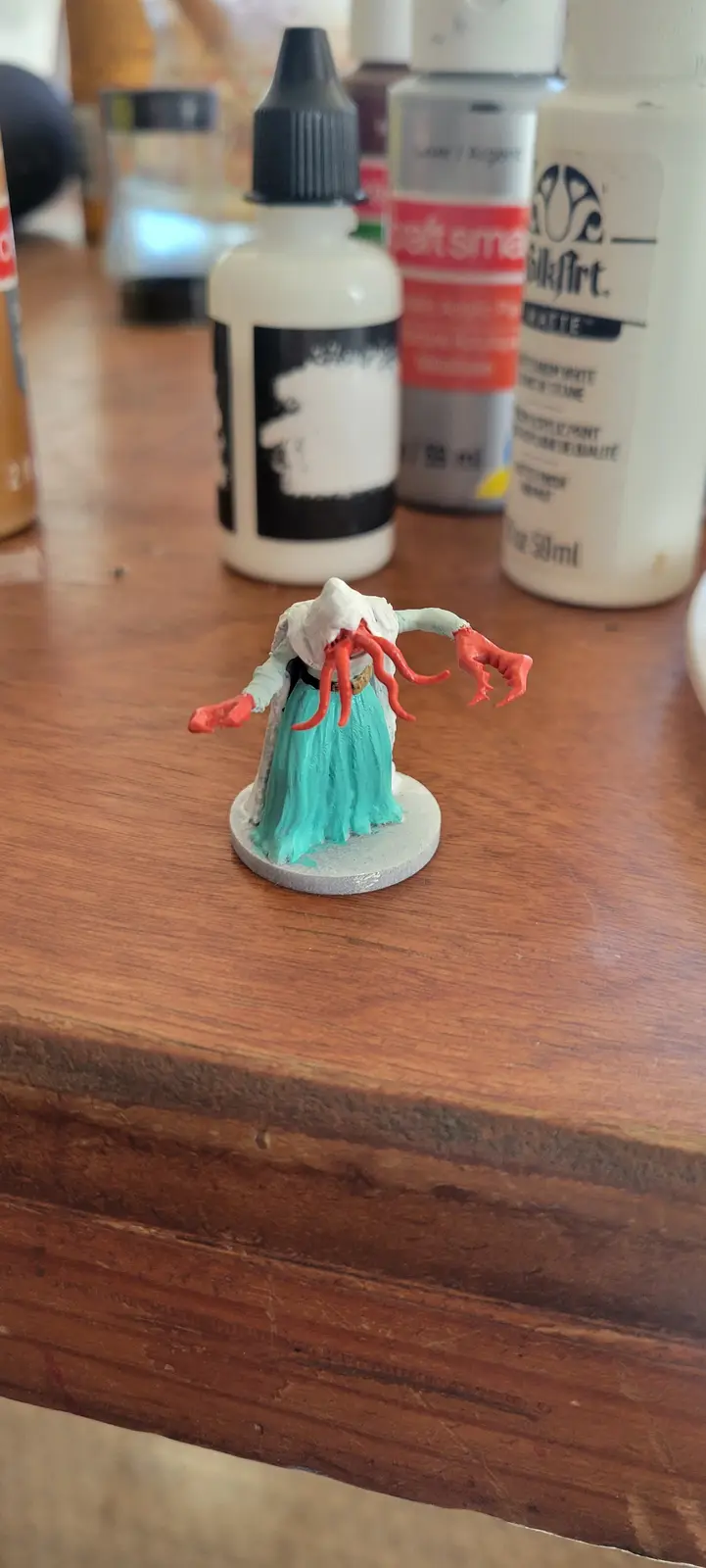 A painted miniature figurine, resembling a humanoid with a white hood, teal skirt, and red tentacles for a face and hands…