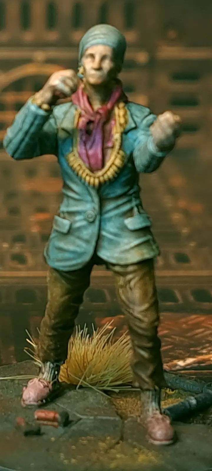 A painted miniature figurine of a person in a blue jacket, pink scarf, and beaded necklace, standing in a fighting stance…
