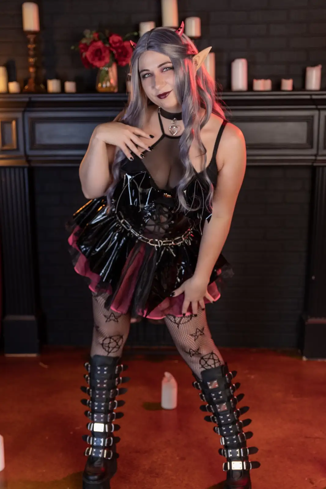 A person with long grey hair, pointed ears, and small red horns poses in a black and pink gothic outfit with fishnet…