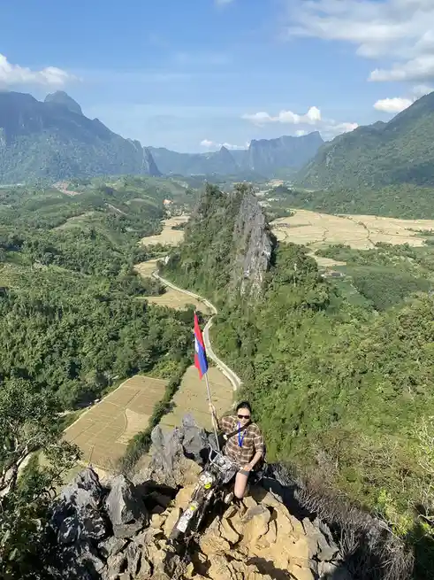 A person wearing sunglasses and a plaid shirt sits atop a rocky cliff edge next to a motorcycle, holding a flag that appears…