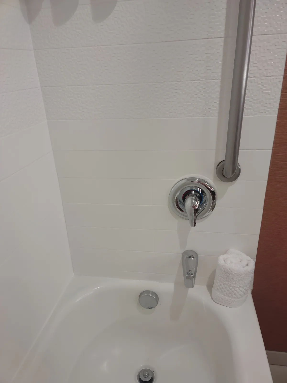 a close-up photo of a white hotel bathtub with chrome fixtures, a rolled white towel on the edge, and a vertical grab bar…