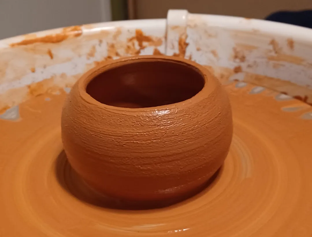 A freshly made, round, orange clay pot sits centered on a spinning pottery wheel, surrounded by wet clay residue on the…