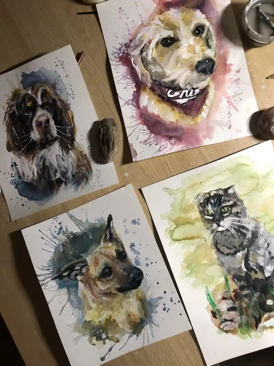 Four watercolor paintings of pets — a spaniel, a smiling dog with a bandana, a German Shepherd, and a cat — are laid out on…