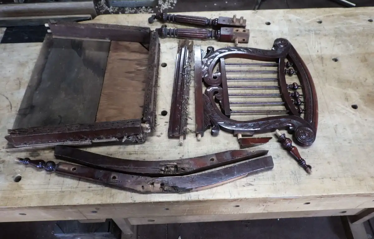A disassembled, worn wooden chair is laid out in pieces on a workbench, showing its broken frame, carved backrest, and…