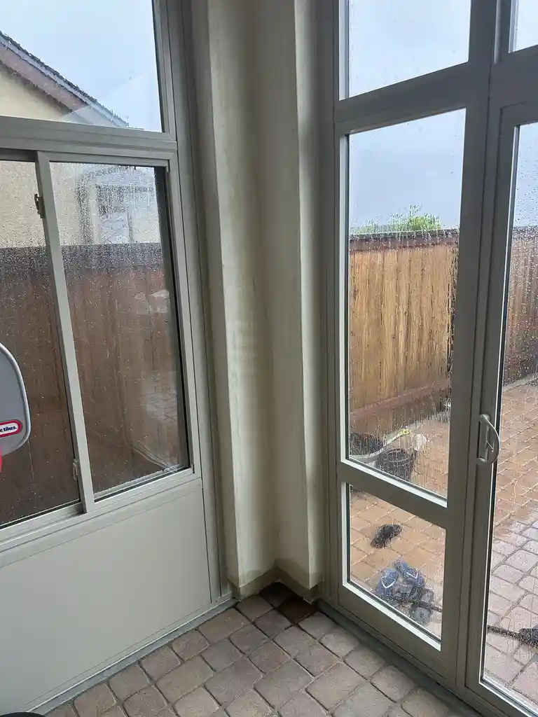A photo shows a rainy day viewed through large glass doors and windows, with water streaks on the glass and a tiled patio…
