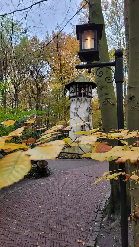 A photo of a park scene with autumn leaves, a brick path, and a small tower in the background. A lit black lantern is in the…
