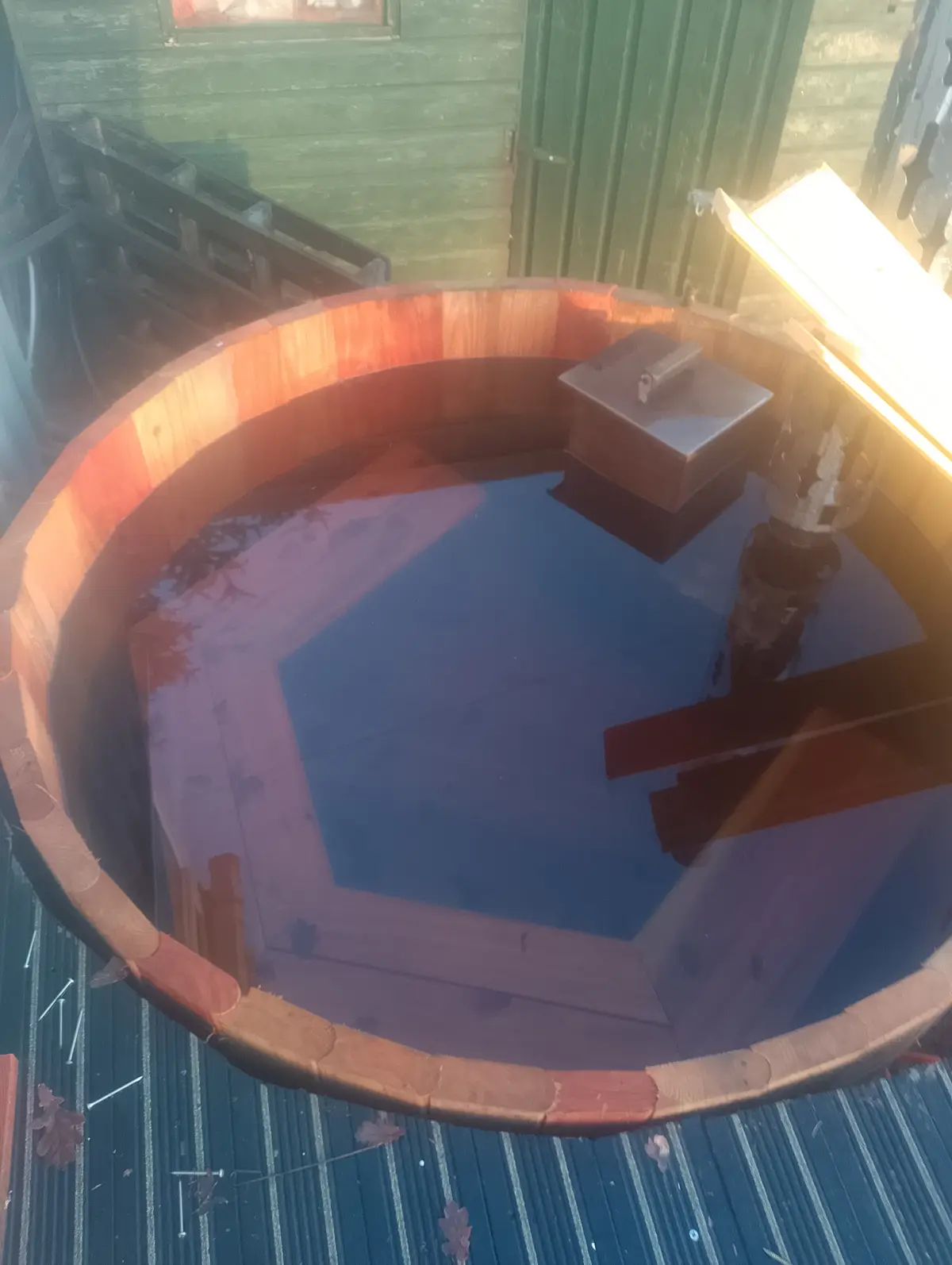 A wooden hot tub filled with water sits outdoors, reflecting a hexagonal wooden deck and a green building in the background…