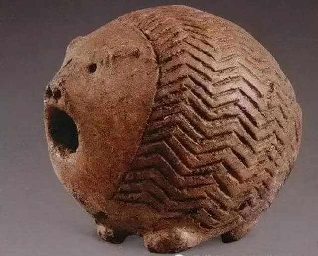 a brown, ancient-looking clay figurine shaped like a pig with a large hole on its side, featuring zigzag patterns etched…