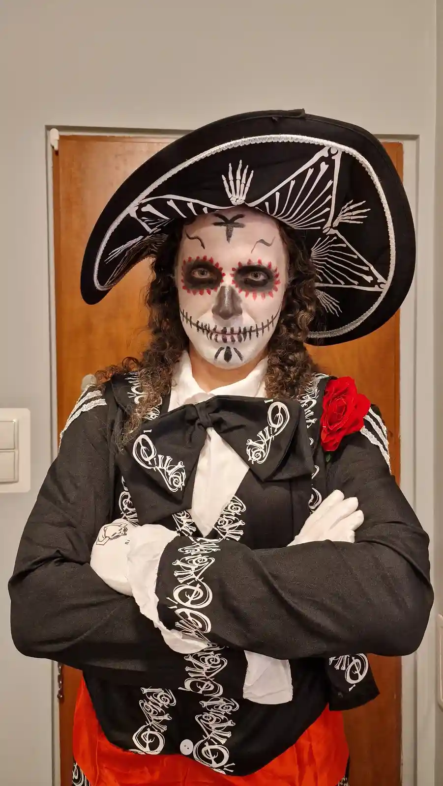 A person dressed as a Day of the Dead skeleton, wearing a large black sombrero with white bone patterns, a black jacket with…