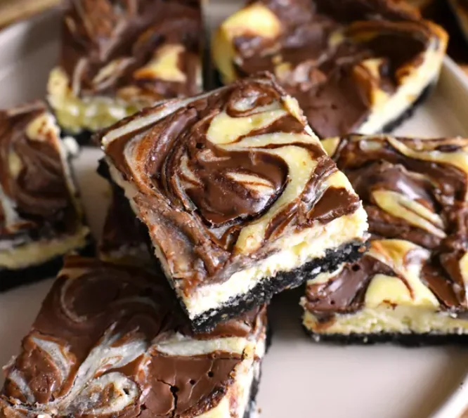 A close-up photo of several square-cut cheesecake bars with a dark chocolate cookie crust, creamy yellow cheesecake filling…