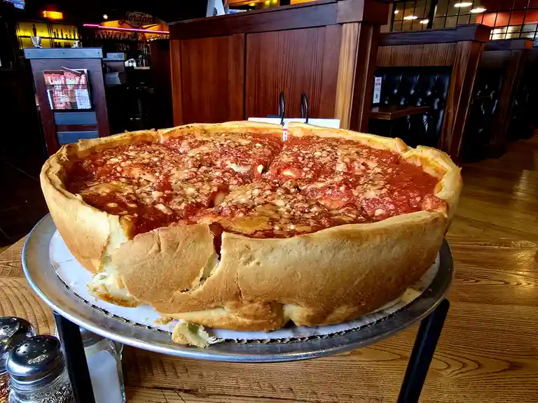 A large, deep-dish pizza with a thick, golden-brown crust sits on a metal tray atop a wooden table in a restaurant. The…