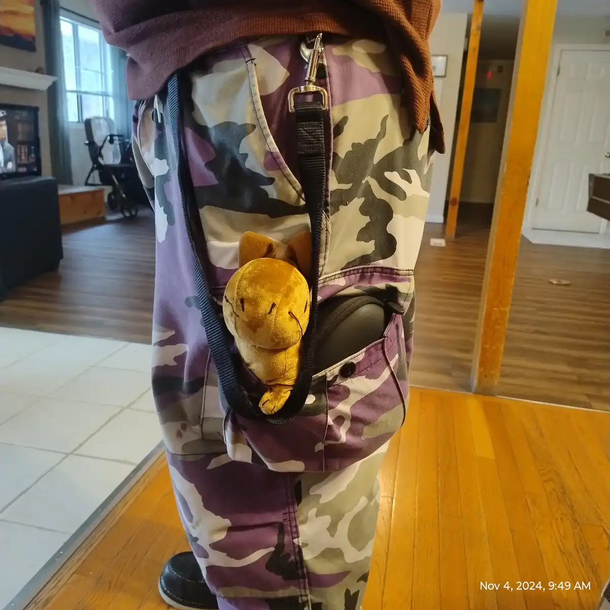 A person wearing purple, green, and beige camouflage pants has a small, yellow plush toy of the Pokémon Diglett peeking out…