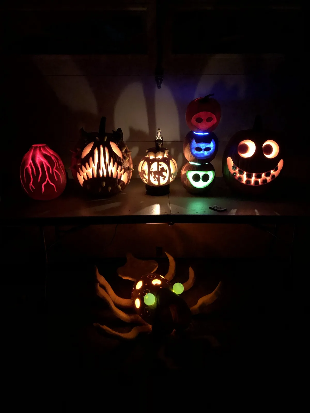A dimly lit photo shows several intricately carved pumpkins with glowing internal lights, arranged on a table and below it…