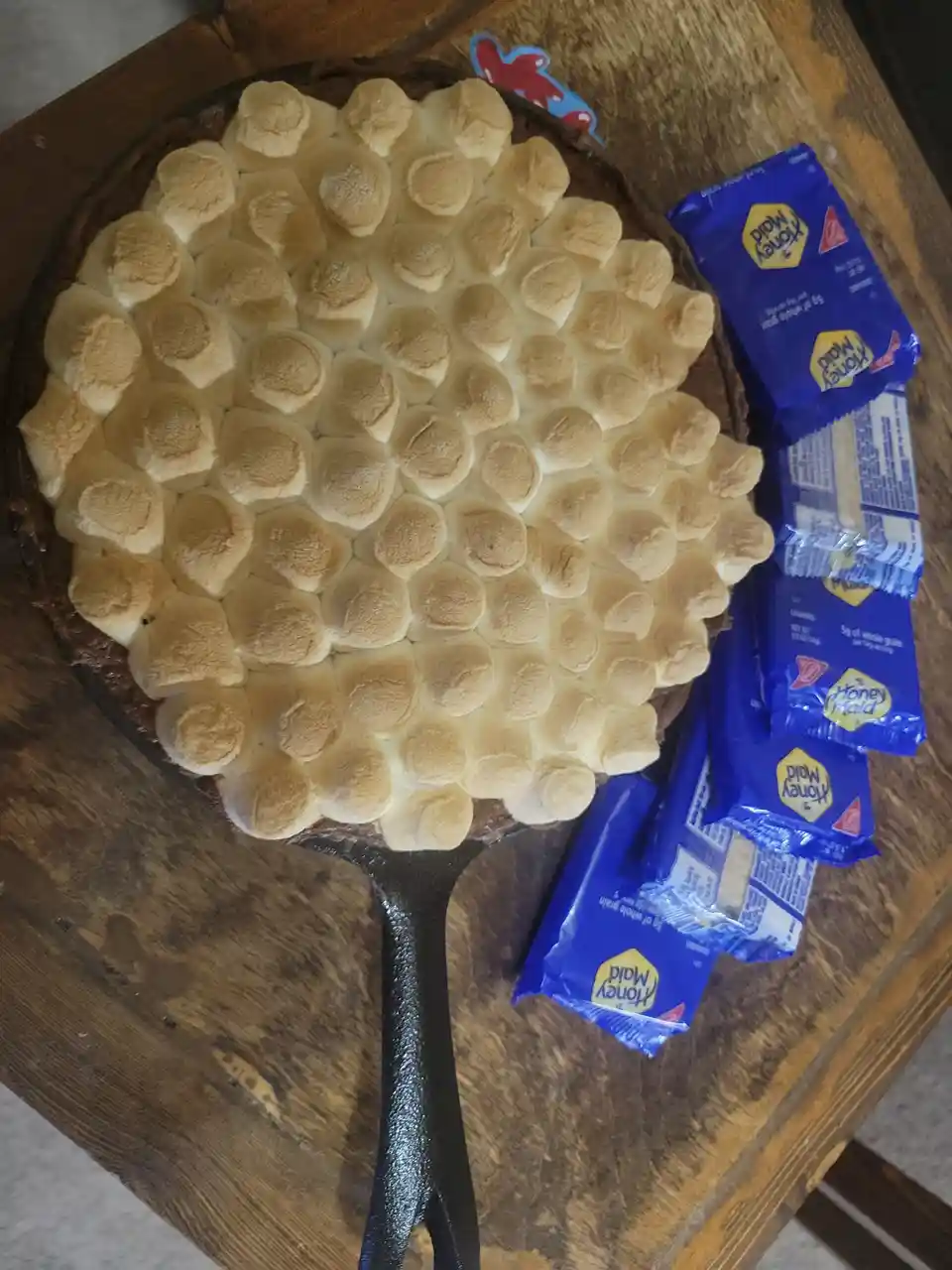 A cast iron skillet filled with a chocolate base and a dense layer of toasted marshmallows sits on a wooden table, next to…