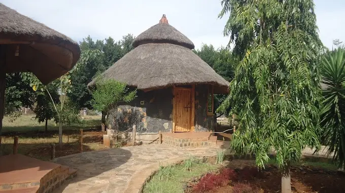 A photo of a thatched-roof hut with stone walls and wooden door, surrounded by trees and a stone path. Overlaid text reads…
