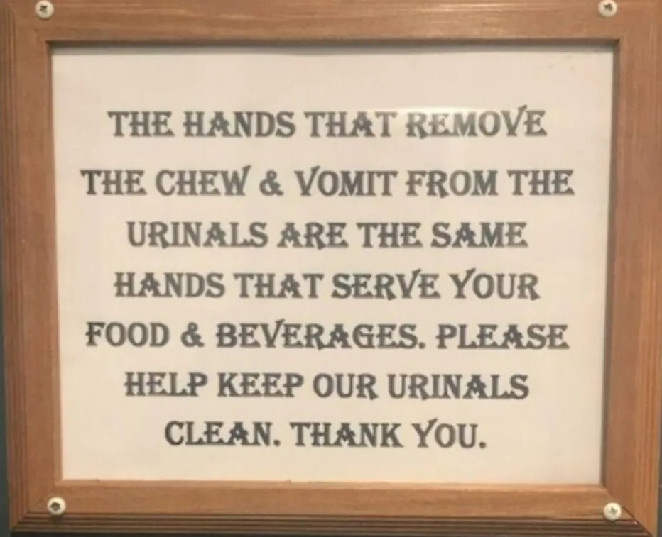 A framed sign with dark text on a light background reads that the same hands cleaning urinals also serve food and drinks…