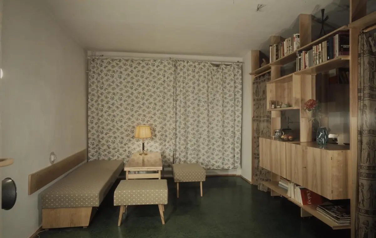 A vintage photograph of a modest, mid-century room with floral curtains, a wooden bench, two patterned stools, a small table…