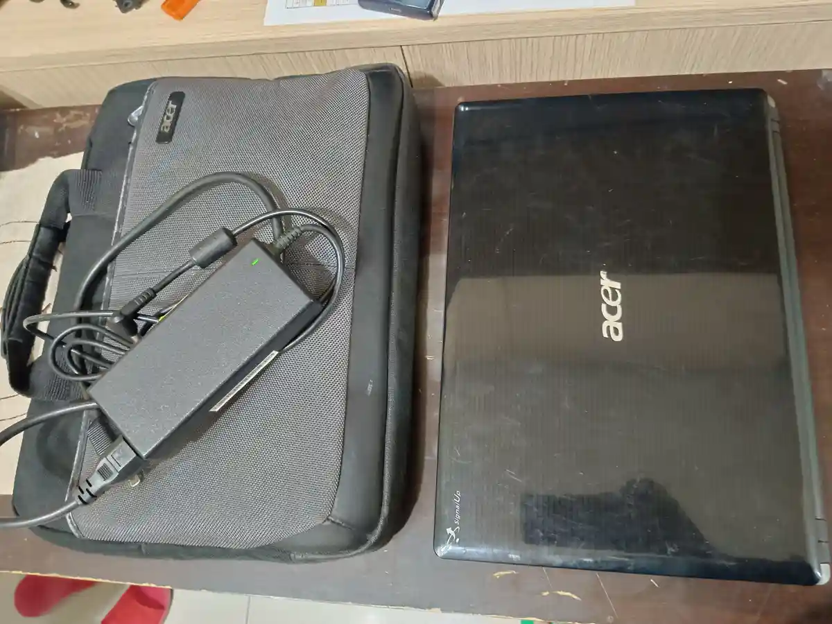 An overhead photo shows a black Acer laptop next to its matching gray fabric carrying case, which has a large, bulky AC…