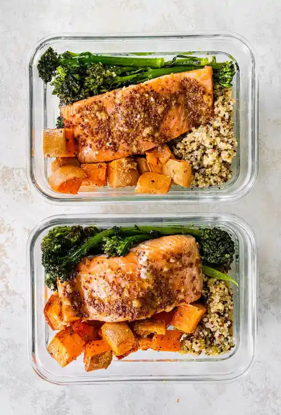 Two identical glass meal prep containers show a healthy meal of salmon, roasted sweet potato cubes, quinoa, and broccolini…