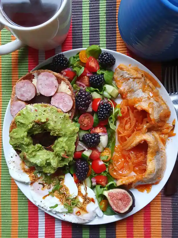 A colorful, overhead shot of a plate of food including avocado toast, bagel with salami, poached eggs, a salad with…