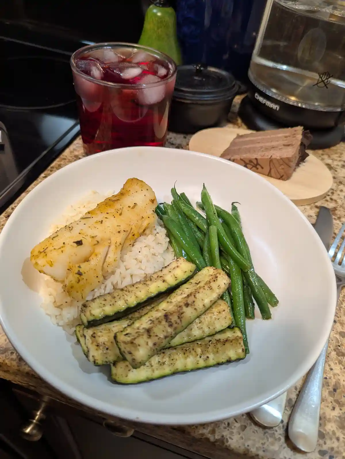 A white plate holds a healthy-looking meal of grilled fish, rice, green beans, and zucchini slices, with a glass of red iced…