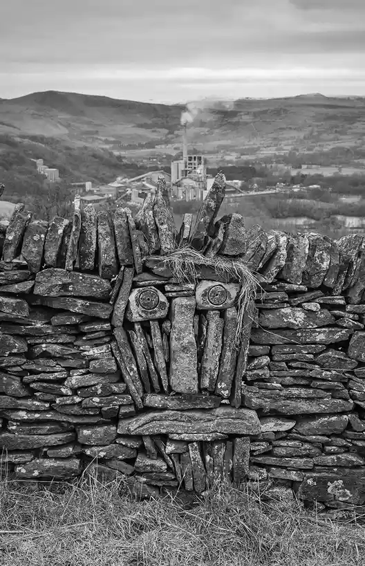 a black and white photograph shows a dry-stone wall in the foreground, where stones have been arranged to resemble a face…